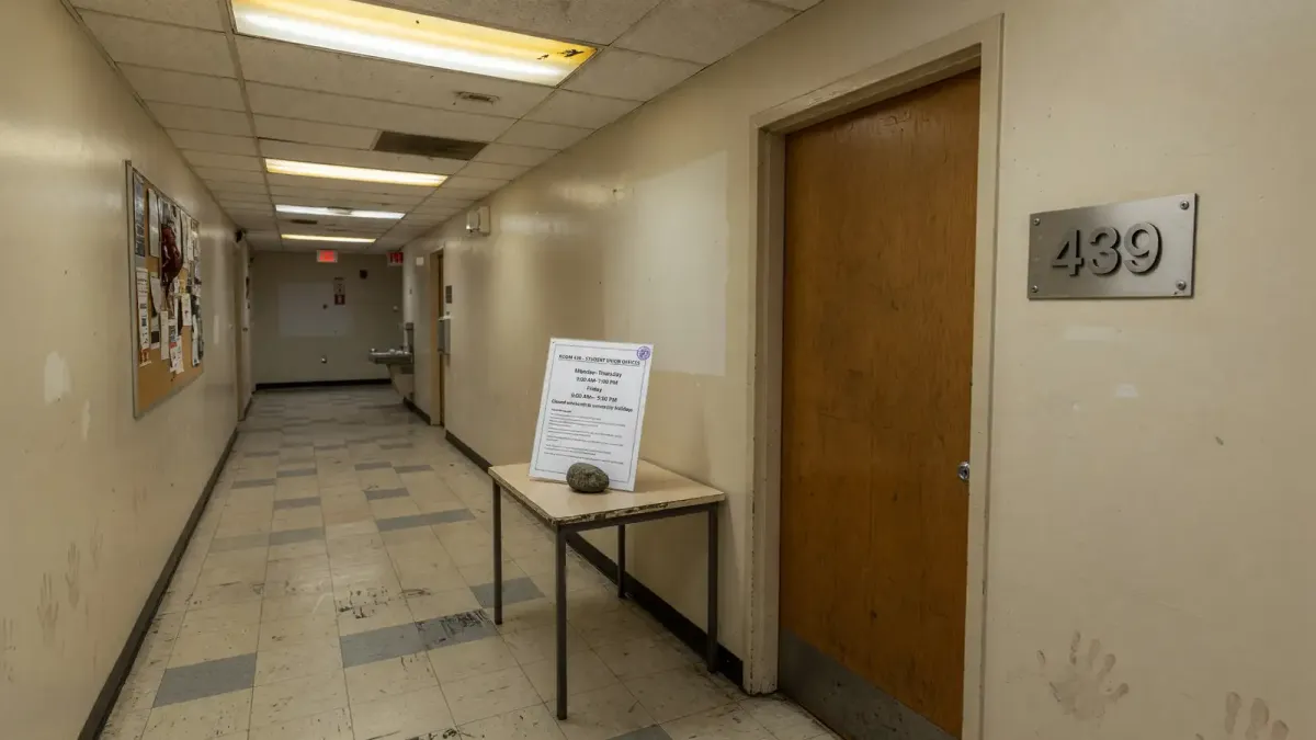 Campus student-union hallway with a suite number plate 439 and a printed resource-center hours sheet on a side table, overhead fluorescent light, no logos