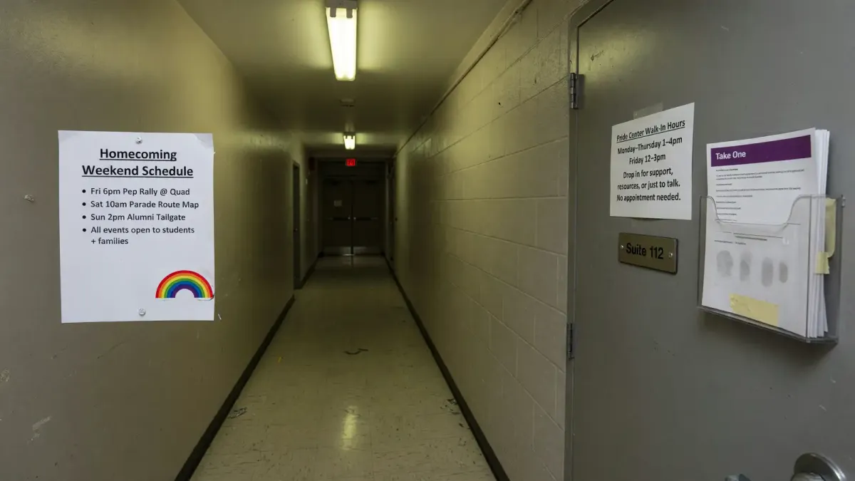Campus union hallway with a paper Homecoming weekend schedule taped beside a closed office door showing printed Pride Center hours, overhead fluorescent light, no logos